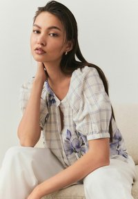 Blusa floreale con maniche a sbuffo, scollo quadrato e un leggero motivo a quadretti in bianco e blu, abbinata a pantaloni bianchi.