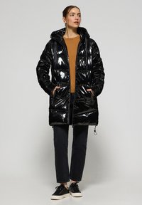 Svart glansig puffjacka med huva, framfickor och logotypdetaljer;parad med en brun topp och mörka jeans; avslappnat fotklädsel.