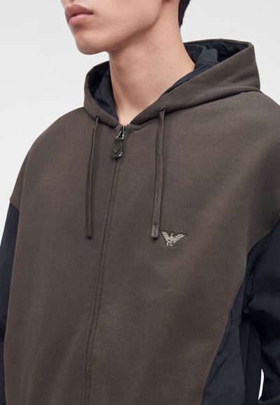 Jeune personne portant un sweat à capuche marron zippé avec une doublure noire à l'intérieur de la capuche et un logo brodé sur la poitrine.
