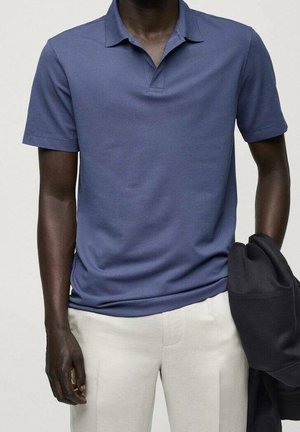 Mann in einem blauen kurzärmeligem Poloshirt und hellbeigen Hosen, der in einer Hand eine gefaltete schwarze Jacke hält.