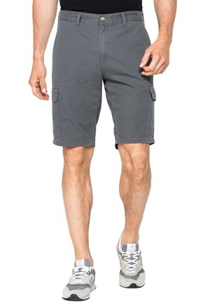 Uomo che indossa pantaloncini cargo grigi e scarpe da ginnastica grigie e bianche, in piedi davanti a uno sfondo bianco.