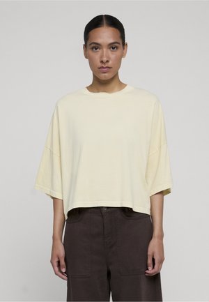 Kvinde med mørkt hår iført en cremefarvet oversized kortærmet bluse og mørke bukser med vide ben, stående mod en ensfarvet lys baggrund.