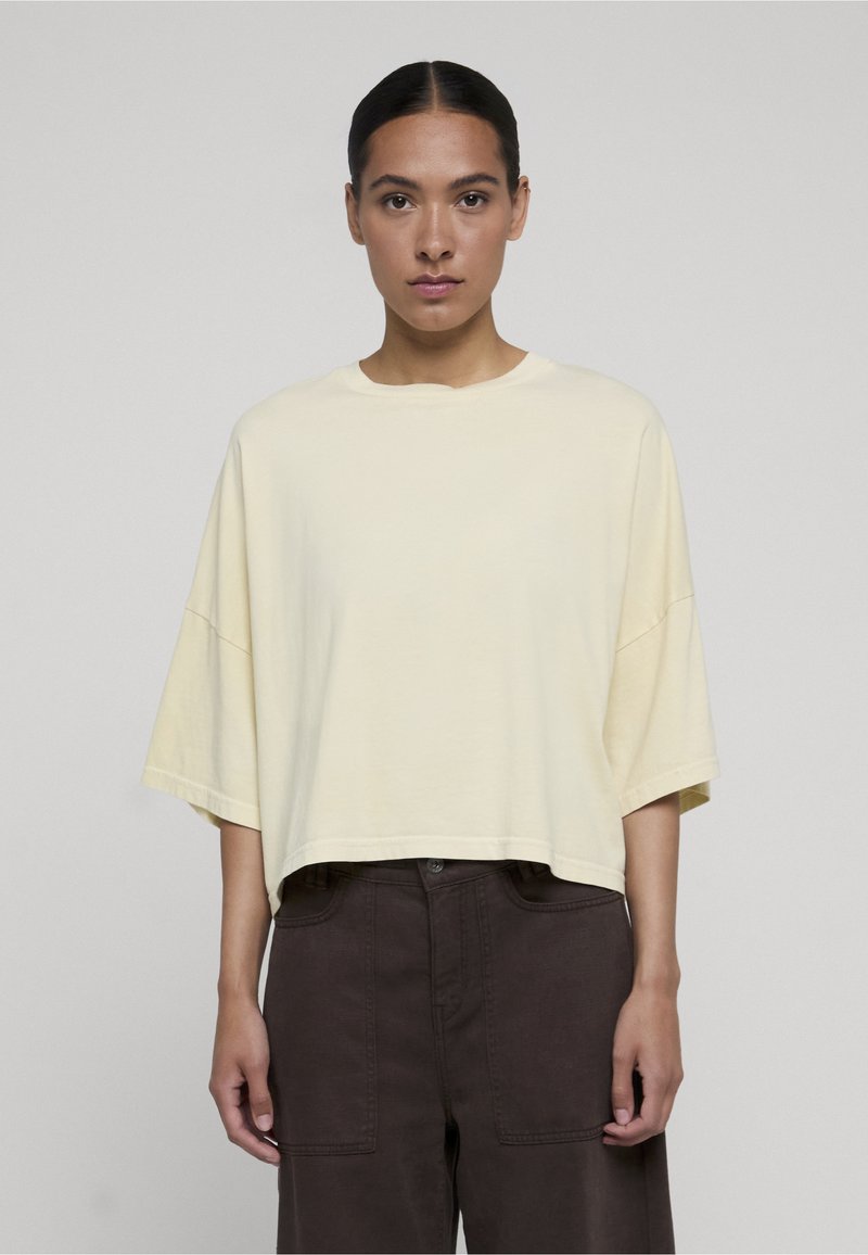 Femme aux cheveux foncés portant un haut crème oversize à manches courtes et un pantalon large foncé, debout devant un fond clair uni.