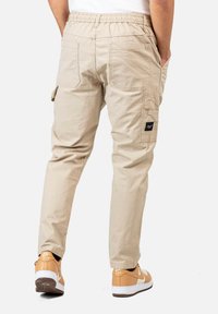 Mann in beigen Cargo-Hosen und tanfarbenen Nike Air Sneakers, der mit den Händen in den Taschen steht, von hinten vor einem weißen Hintergrund gezeigt.