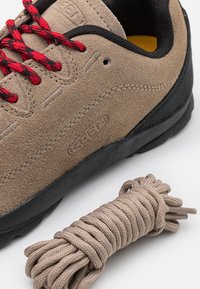 Chaussure en daim brun avec semelle noire et lacets rouges, à côté d'un lacet beige enroulé sur un fond blanc.