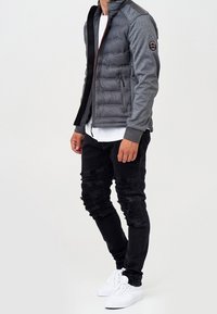 Mann trägt eine graue gesteppte Jacke mit Stoffärmeln, weißem Hemd, schwarzen zerrissenen Jeans und weißen Turnschuhen, steht vor einem schlichten Hintergrund.