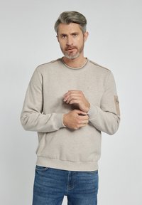 Jasnobeżowy sweter z okrągłym dekoltem, ściągaczami na mankietach i detalami szycia na ramionach. Posiada teksturowaną łatkę na lewym rękawie.