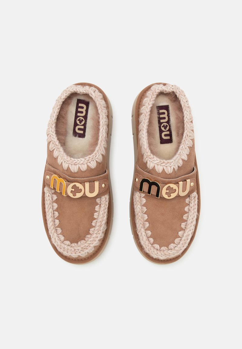 Een paar bruine suède mocassinslippers met crèmekleurige gebreide rand en een metalen logo van het merk op het bandje, gezien van bovenaf op een witte achtergrond.