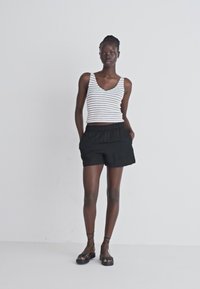 Débardeur rayé blanc et marine, shorts noirs à taille élastique, design minimaliste, avec des sandales noires à lanières sur un fond neutre.