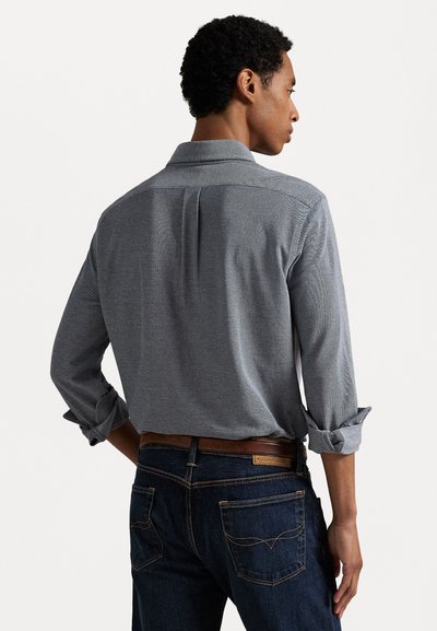 Gray long-sleeve shirt with a button-down collar and rolled cuffs, featuring a back pleat. Worn with dark jeans and a brown belt.