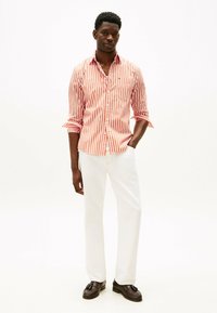 Pantaloni bianchi da uomo abbinati a una camicia a righe rosse e bianche con bottoni. La camicia presenta una tasca e le maniche arrotolate. Mocassini scuri completano l'outfit.
