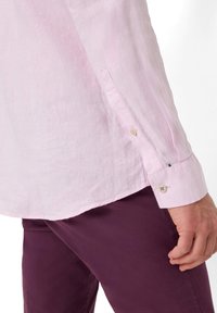 Camicia in lino rosa chiaro con maniche lunghe, polsini abbottonati, orlo curvo, abbinata a pantaloni viola scuro, con una superficie liscia.