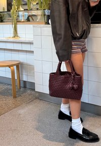 Borsa intrecciata bordeaux con una forma strutturata, finitura testurizzata e due pompon decorativi; abbinata a scarpe nere e calze bianche.
