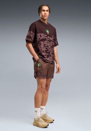 Jeune homme aux cheveux tressés portant un t-shirt camouflage marron et un short, des chaussettes blanches Puma et des baskets beiges, debout sur fond gris.