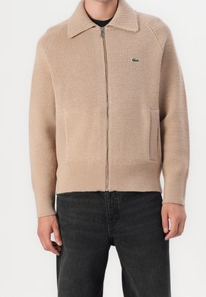 Homme portant un pull en maille beige zippé avec col et logo Lacoste, assorti à un jean noir, debout devant un fond blanc.