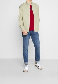 Světle zelená košile s dlouhým rukávem a knoflíky, pod ní červené tričko se šedým pruhem, doplněná modrými džíny a bílými teniskami.