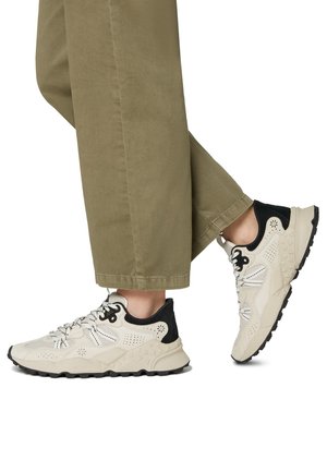 Beige sportsko med sorte accenter, mesh- og syntetisk overdel, polstret krave, gummisål og reflekterende detaljer nær snørebåndene.