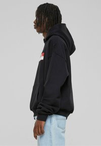 Black hoodie with a front pocket, loose fit, and drawstring hood. Features a red logo patch on the chest. Worn with light blue jeans.