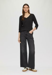 Zwart langemouwen V-hals top gecombineerd met wijde donkere jeans en witte sneakers met bruine zolen. Eenvoudig, modern ontwerp en lichtgewicht stof.