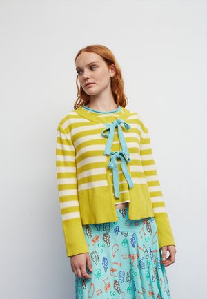 Femme aux cheveux roux portant un cardigan rayé jaune et blanc avec des nœuds bleus sur une jupe turquoise à motifs, debout sur un fond blanc.