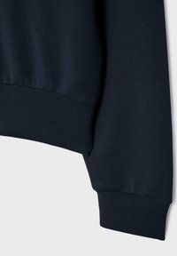 Gros plan sur la manche d'un sweat-shirt bleu marine avec un poignet et un ourlet côtelés sur un fond clair uni.