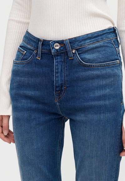 Μπλε τζιν από denim μπροστινή όψη με κουμπί και φερμουάρ, φορεμένο με λευκό ριμπ μακρυμάνικο τοπ.