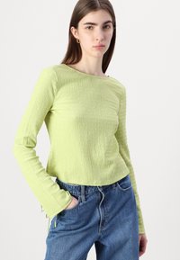 Limegrønn teksturert langermet topp med båtutringning og justerbare knytebånd ved mansjettene, kombinert med løstsittende denimjeans.