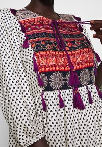 Bohemisk stil klänning med svart och vit liten pilmönster, livfullt blommigt panel, lila tofsar och flätad knytning vid halsringningen på mörk hudton.