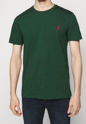 Man draagt een donker groen T-shirt met korte mouwen en een klein rood geborduurd logo op de linkerborst, gecombineerd met donkerblauwe spijkerbroek.