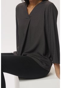 Tmavě šedá blůza s výstřihem do V, ozdobená texturovaným tečkovaným vzorem. Volný střih, 3/4 rukávy a hladký materiál. Nosič má na sobě černé kalhoty.