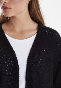 Černý pletený cardigan s otevřeným vzorem a žebrovanými okraji, nošený přes jednoduchou bílou košili, s uvolněným střihem a výstřihem do V.