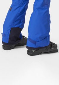 Blauwe waterdichte broek met donkerblauwe accenten en klittenbandsluitingen, gecombineerd met zwarte buitenschoenen met een gestructureerd oppervlak en een ondersteunend ontwerp.