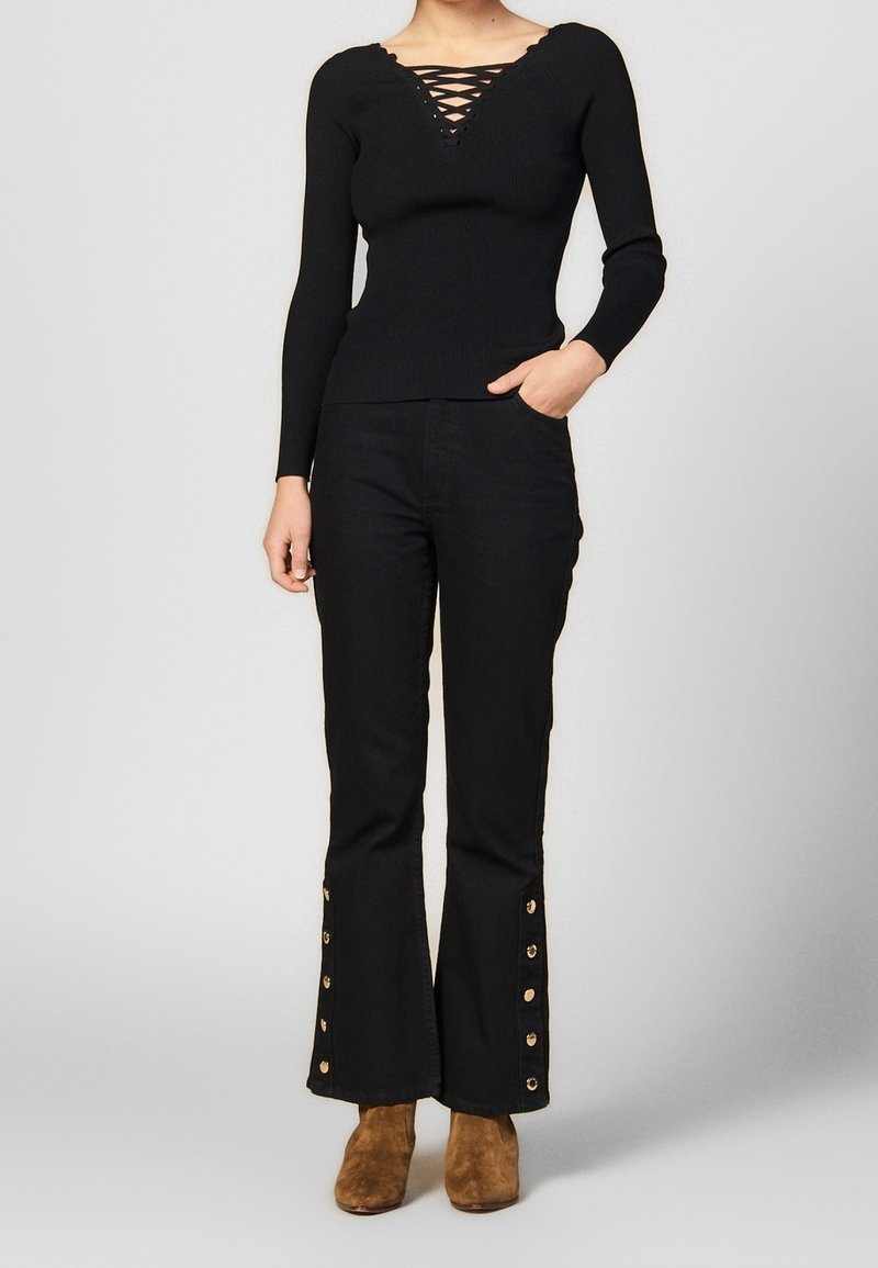 Femme portant un haut noir à manches longues avec un col croisé, associé à un pantalon noir avec des boutons dorés le long des poignets évasés et des bottines marron.