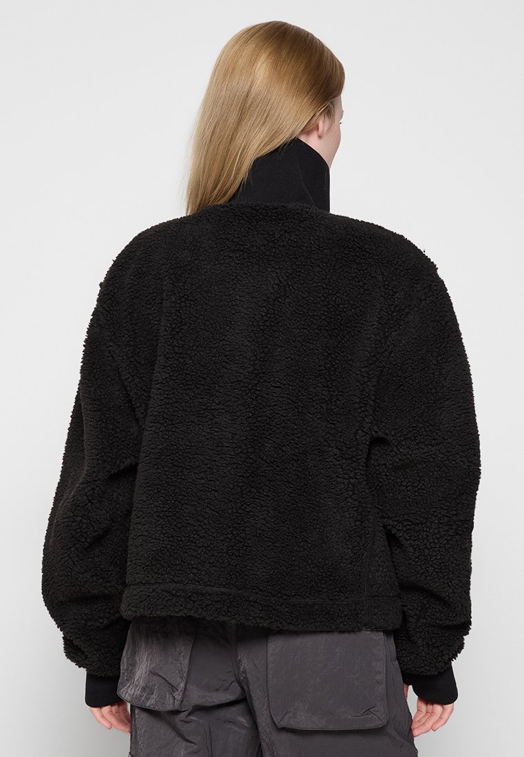 Veste polaire noire surdimensionnée avec une finition texturée et confortable. Caractéristiques : col haut et épaules tombantes. Associée à un pantalon cargo gris.