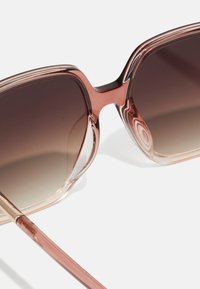 Sunglasses with a translucent rose-colored frame, gradient brown lenses, and slender arms featuring a slight gold accent at the hinge.