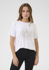 T-shirt en coton blanc avec un col rond et des manches courtes ; présente un motif graphique doré central complexe avec des lignes rayonnantes.