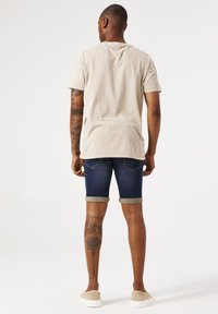 Camiseta beige de manga corta, pantalones cortos de mezclilla azul doblados en el dobladillo, con un tatuaje en la pierna y calzado slip-on beige claro.