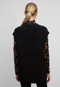 Gilet lavorato a maglia grigio scuro con texture a righe, caratterizzato da ampie aperture per le braccia e un colletto alto, indossato sopra una maglietta nera a maniche lunghe con motivo floreale.