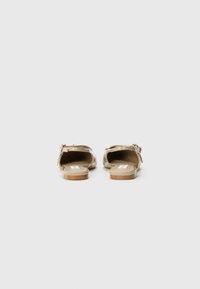Beige open-back sandals with a smooth patent finish, flat soles, and adjustable strap featuring a small silver buckle.