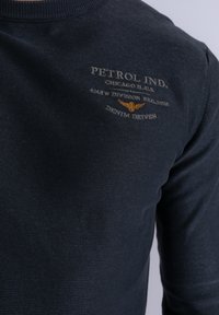 Ciemnoniebieski sweter z prążkowanym kołnierzem, tkaną teksturą oraz złotą haftowaną grafiką z napisem "Petrol Ind." i "Denim Driven." Godne uwagi detale to logo i wzór.