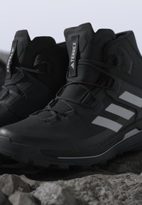 Sorte Adidas Terrex vandrestøvler med reflekterende striber og forstærkede såler placeret på klippegrund udendørs.