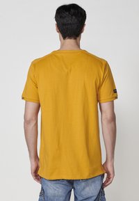 Geltona trumpomis rankovėmis marškinėliai, apvaliu kaklu ir vienodai audiniu. Išskirtinis dryžuotas detalė ant rankovės. Rodyti iš nugaros.