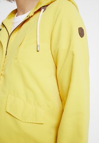 Gul hættejakke med lynlås, forlommes og dekorativ brun lap på ærmet. Let og glat tekstur.