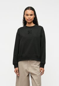 Μαύρο φούτερ με στρογγυλή λαιμόκοψη, φουσκωτά μανίκια και μικρό ανάγλυφο λογότυπο στο στήθος. Κατασκευασμένο από μαλακό, υφασμάτινο υλικό.