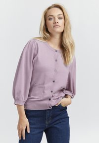 Ljuslila kofta med rund halsringning, knap stängning, två framfickor och puffade ärmar, tillverkad av ett mjukt, texturerat material.