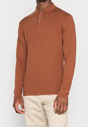 Mand iført en rustfarvet langærmet polo med tre knapper, kombineret med lyse beige bukser mod en ensfarvet baggrund.
