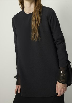 Frau trägt ein langes schwarzes Kleid mit durchsichtigen Bündchen und glänzenden schwarzen Armbändern, steht vor einem schlichten hellen Hintergrund.