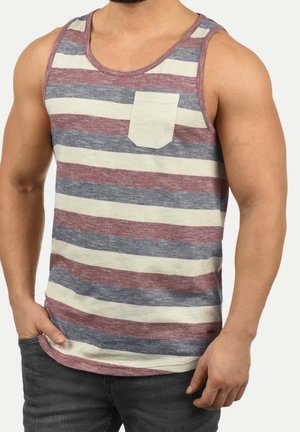 Homme portant un débardeur à rayures horizontales beige, bordeaux et gris, avec une petite poche poitrine beige, assorti à un jean gris foncé.