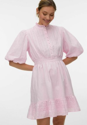 Žena v svetloružových šatách s nafúknutými rukávmi, zapínanými na gombíky, s detailmi z dierkovanej čipky a volánikmi na leme, usmieva sa a pozerá dole.