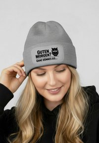 Gray beanie with ribbed texture, featuring a rectangular patch with black owl graphic and German text: "GUTEN MORGEN? GANZ DÜNNES EIS..."